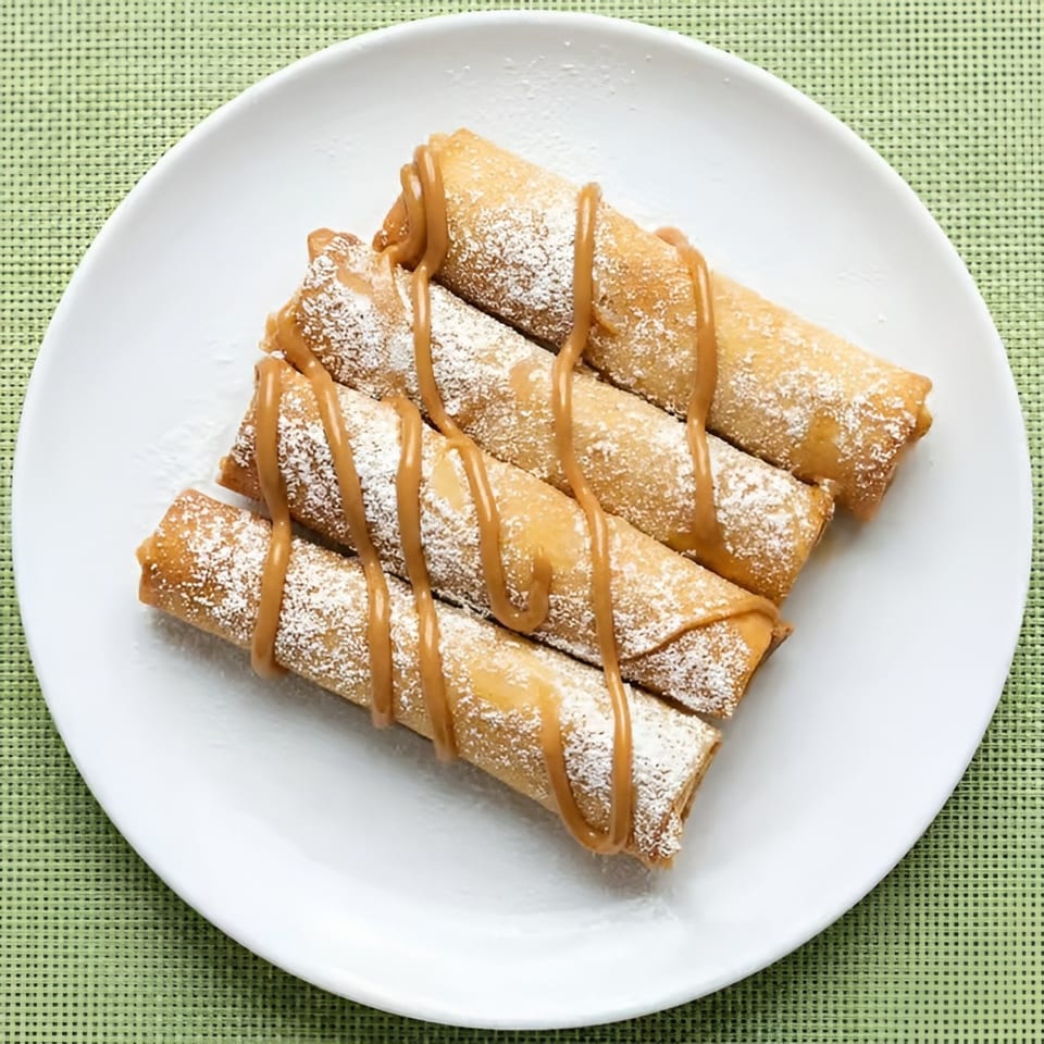 Four golden-brown dessert spring rolls, drizzled with caramel and dusted with powdered sugar on a white plate.
